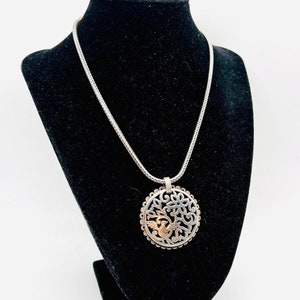 May include: A silver necklace with a round pendant featuring a delicate floral design and a small bird. The pendant is made of silver and has a textured finish.