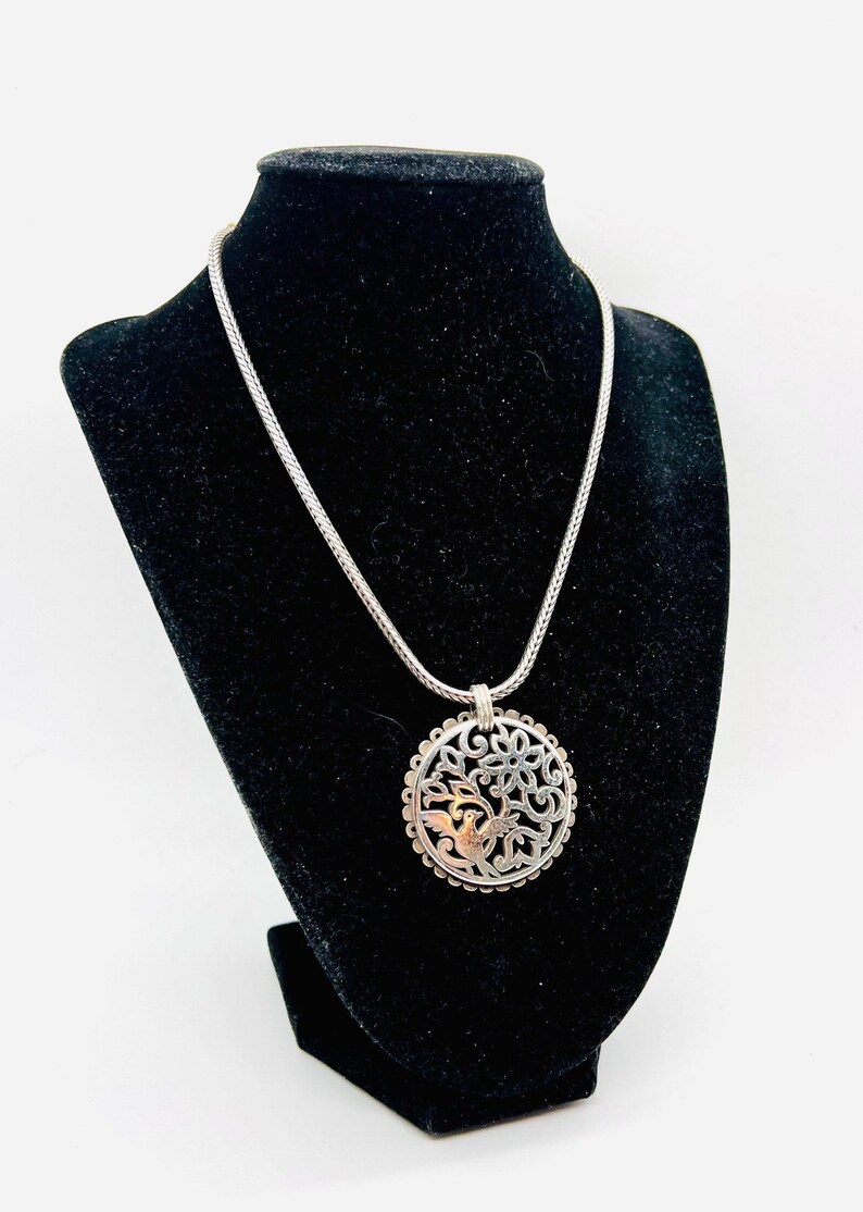 May include: A silver necklace with a round pendant featuring a delicate floral design and a small bird. The pendant is made of silver and has a textured finish.