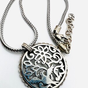 May include: Silver necklace with a round pendant featuring a delicate filigree design of a bird in a tree. The chain is a silver box chain.