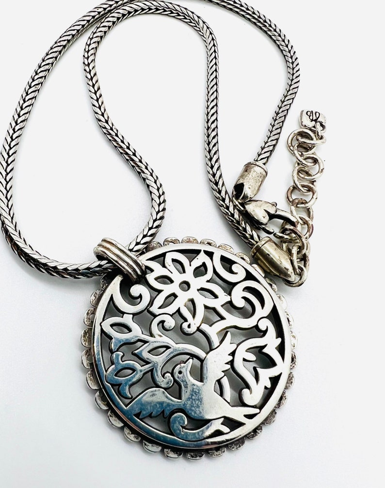 May include: Silver necklace with a round pendant featuring a delicate filigree design of a bird in a tree. The chain is a silver box chain.