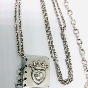 May include: A silver necklace with a charm that looks like a small book with a heart and crown design. The chain is a double chain with a lobster clasp.