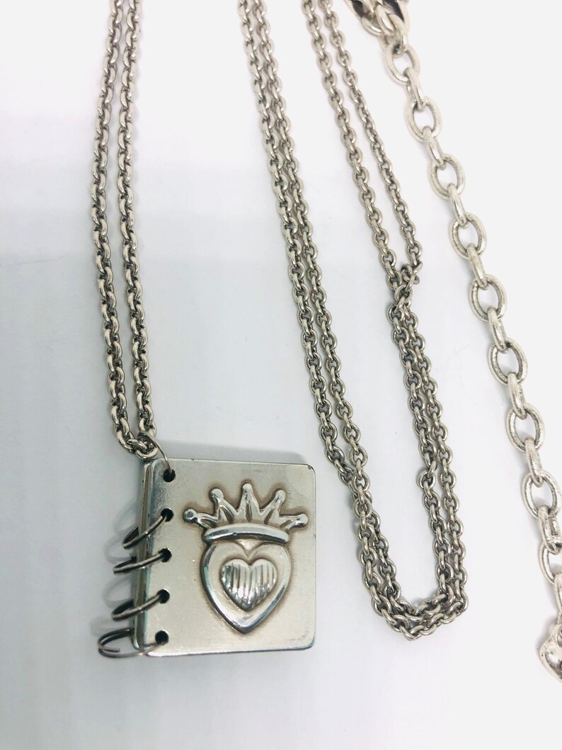 May include: A silver necklace with a charm that looks like a small book with a heart and crown design. The chain is a double chain with a lobster clasp.
