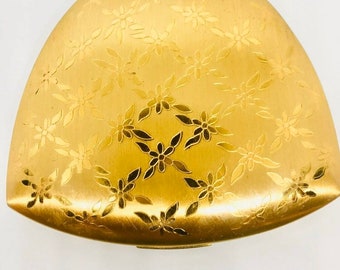 May include: A gold-colored compact with a floral design. The compact is closed and has a rounded, slightly curved shape.