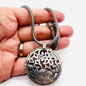 May include: A silver chain necklace with a round pendant featuring a cutout design of a tree with birds. The pendant has a detailed floral pattern and a textured border.