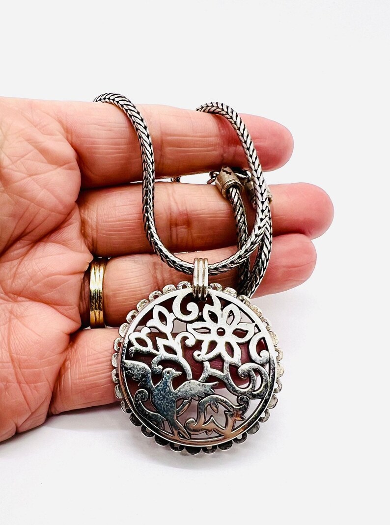 May include: A silver chain necklace with a round pendant featuring a cutout design of a tree with birds. The pendant has a detailed floral pattern and a textured border.