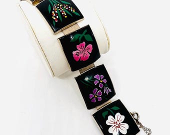 May include: A silver bracelet with black enamel squares featuring hand-painted floral designs in pink, purple, white, and green.
