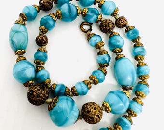 May include: A vintage necklace with blue glass beads and gold metal accents. The beads are oval shaped and have a marbled effect. The necklace is strung on a thin cord.