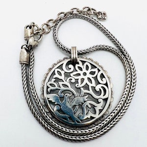 May include: A silver necklace with a delicate chain and a pendant featuring a circular cutout design of a bird in a tree with a flower. The pendant is detailed with intricate swirls and floral motifs.