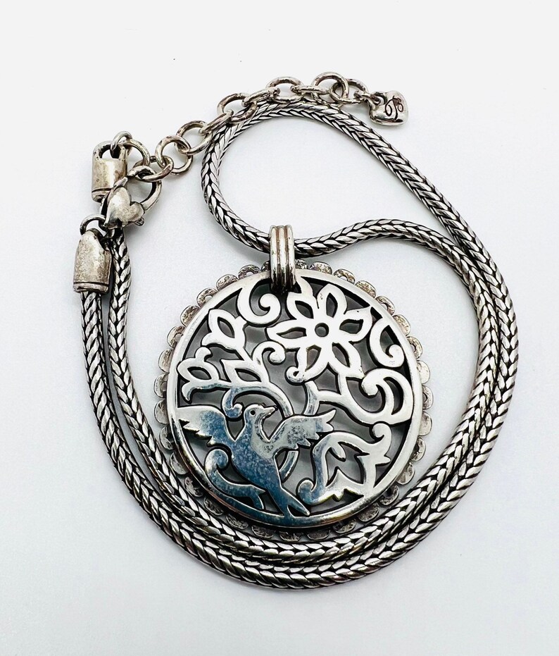 May include: A silver necklace with a delicate chain and a pendant featuring a circular cutout design of a bird in a tree with a flower. The pendant is detailed with intricate swirls and floral motifs.