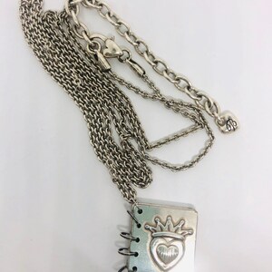 May include: A silver necklace with a charm that looks like a small notebook with a heart and crown design. The charm has a ring binder style opening.