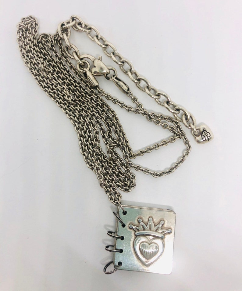 May include: A silver necklace with a charm that looks like a small notebook with a heart and crown design. The charm has a ring binder style opening.