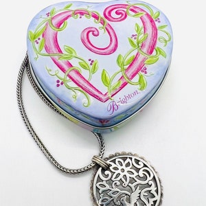 May include: A heart-shaped tin box with a pink and green floral design and the word "Brighton" on the bottom. The box is attached to a silver chain with a silver pendant featuring a bird in a tree design.