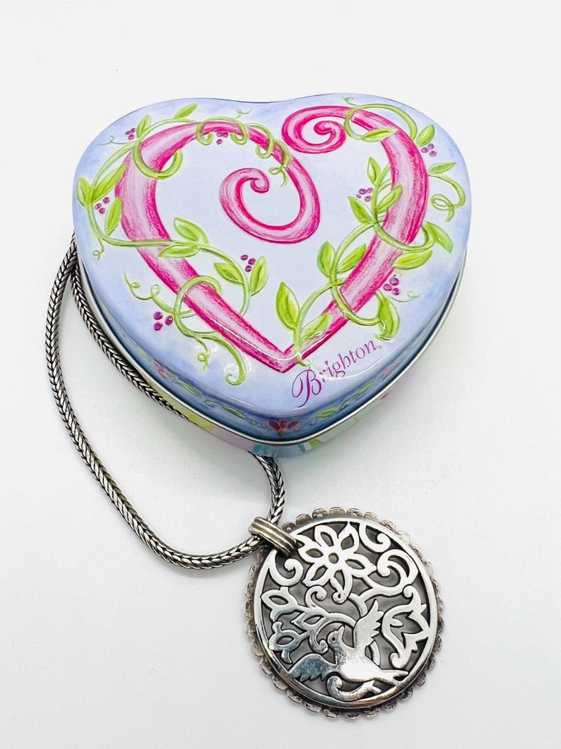 May include: A heart-shaped tin box with a pink and green floral design and the word "Brighton" on the bottom. The box is attached to a silver chain with a silver pendant featuring a bird in a tree design.