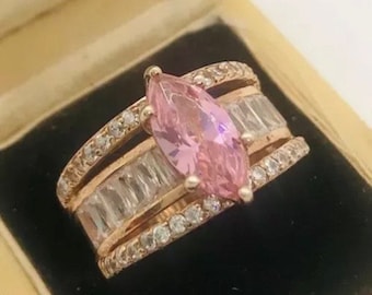 May include: A rose gold ring with a large pink marquise-shaped gemstone surrounded by smaller clear round gemstones. The ring is set with three rows of smaller clear gemstones.