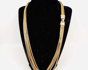 May include: A gold-toned necklace with multiple strands, displayed on a black mannequin. The necklace features a rectangular clasp and hangs in a layered design. The jewelry is set against a white background.