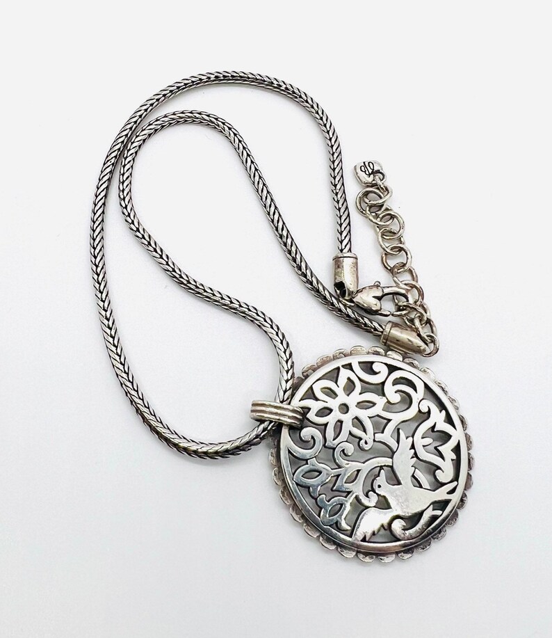 May include: A silver necklace with a round pendant featuring a delicate, cutout design of a bird and floral motifs.