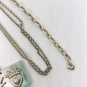 May include: A silver necklace featuring a charm in the shape of a small book with a heart and crown on the cover. The necklace has two chains, one is a cable chain and the other is a curb chain. The charm has a small heart-shaped tag with the letter "B" on it.