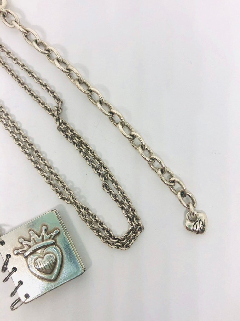 May include: A silver necklace featuring a charm in the shape of a small book with a heart and crown on the cover. The necklace has two chains, one is a cable chain and the other is a curb chain. The charm has a small heart-shaped tag with the letter "B" on it.