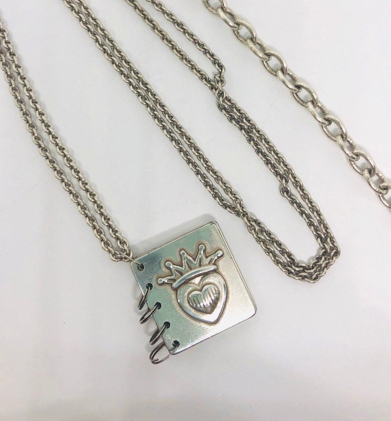 May include: A silver necklace with a silver charm in the shape of a small book with a heart and crown design. The necklace has two chains.