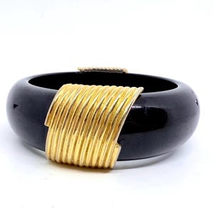May include: A black, wide bangle bracelet with a gold-tone metal band. The gold band wraps around the center of the bracelet, creating a striking contrast. The bracelet is set against a white background.