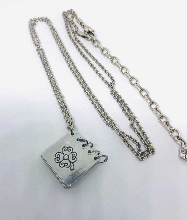 May include: A silver necklace featuring a silver charm in the shape of a small book with a four-leaf clover design. The chain is a delicate, double-strand chain.