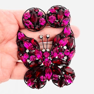 May include: A large, pink and red rhinestone butterfly brooch with a black metal frame. The brooch is accented with clear rhinestones and has a pin back.