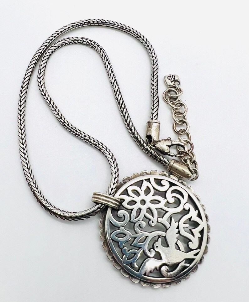May include: A silver necklace with a delicate chain and a pendant featuring a circular design with a bird and floral motifs.