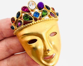 May include: Gold-toned metal mask brooch with a crown of colorful rhinestones. The mask has a smiling expression and is decorated with a variety of colored gems.