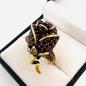May include: A gold-tone ring with a red rose design. The rose is made of small red crystals and has a green stem.