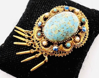 May include: A gold-tone brooch with a large blue and white oval cabochon stone surrounded by smaller blue and white stones. The brooch has a dangling fringe of gold chains and spikes.