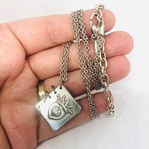 May include: A silver necklace with a square pendant featuring a heart with a crown on top. The chain is a delicate, layered design.