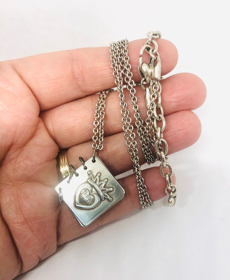 May include: A silver necklace with a square pendant featuring a heart with a crown on top. The chain is a delicate, layered design.