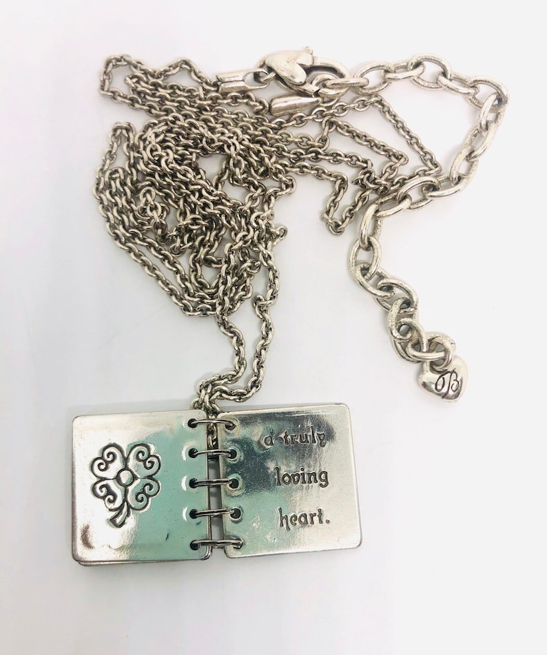 May include: Silver necklace with a charm that looks like a small book with a four-leaf clover on the front and the words "a truly loving heart" on the back.
