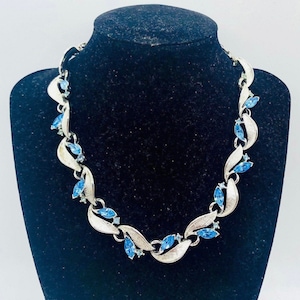 May include: Silver-toned necklace with a leaf design, adorned with marquise-cut blue gemstones. The necklace is displayed on a black mannequin, showcasing its intricate details and elegant style. A classic piece of jewelry.
