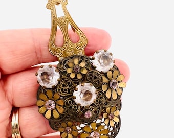 May include: Vintage brass-toned metal brooch with a floral design. The brooch features clear and pink crystal accents, and filigree detailing. The clasp is a decorative, ornate design. The brooch is held in a hand.