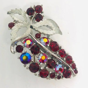 May include: A silver tone brooch with a leaf design and red and iridescent rhinestones.