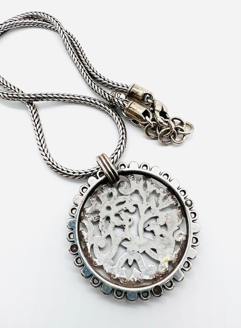 May include: A silver necklace with a round pendant featuring a detailed, intricate design. The pendant is made of silver and has a delicate, filigree pattern.