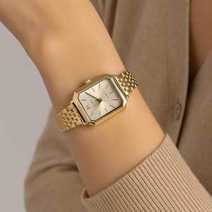 May include: A gold wristwatch with a rectangular face and a matching link bracelet. The watch face is light-coloured with black hands and markers. The watch is worn on a wrist, with a neutral-toned jumper in the background.
