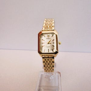 Puede incluir: Un reloj de pulsera dorado con una esfera rectangular y una correa de eslabones a juego. La esfera del reloj es blanca con marcadores negros y la marca "XIMILI". El reloj se exhibe en un soporte de pl&aacute;stico transparente.