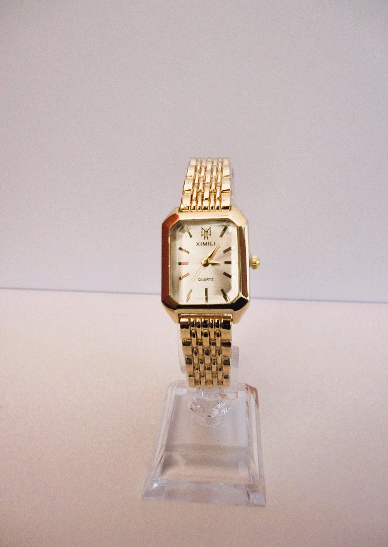Puede incluir: Un reloj de pulsera dorado con una esfera rectangular y una correa de eslabones a juego. La esfera del reloj es blanca con marcadores negros y la marca "XIMILI". El reloj se exhibe en un soporte de pl&aacute;stico transparente.