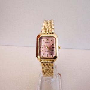 Puede incluir: Un reloj de cuarzo rectangular dorado con esfera rosa y correa dorada a juego. La esfera del reloj tiene agujas e indicadores dorados. El reloj se exhibe sobre un soporte de pl&aacute;stico transparente. La marca "XIMILI" es visible.