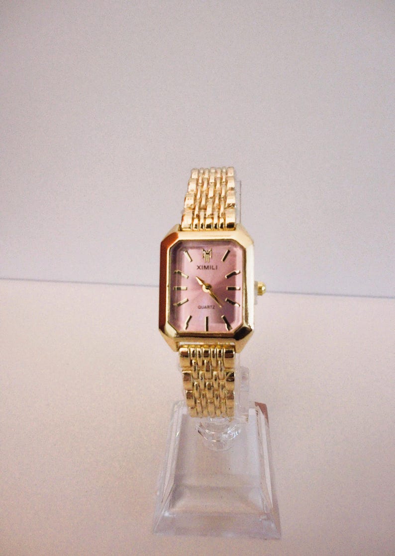 Puede incluir: Un reloj de cuarzo rectangular dorado con esfera rosa y correa dorada a juego. La esfera del reloj tiene agujas e indicadores dorados. El reloj se exhibe sobre un soporte de pl&aacute;stico transparente. La marca "XIMILI" es visible.