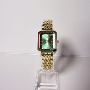Puede incluir: Un reloj de pulsera dorado con una esfera verde rectangular y una correa dorada a juego. El reloj se exhibe sobre un soporte transparente. La esfera del reloj tiene agujas e indicadores de hora dorados.