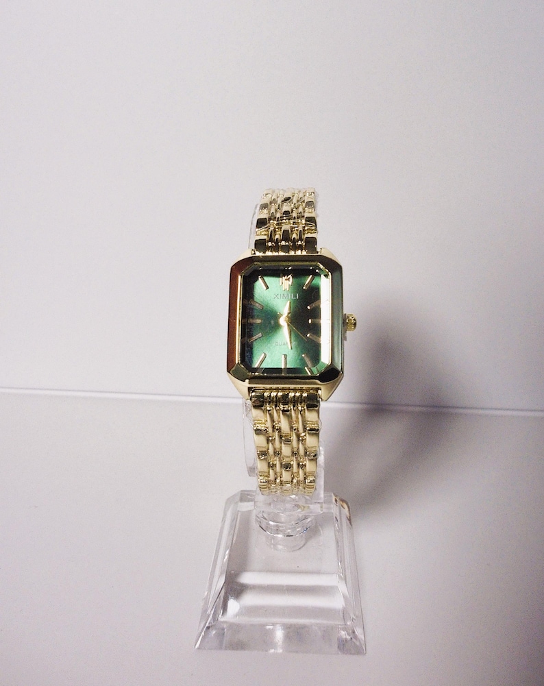 Puede incluir: Un reloj de pulsera dorado con una esfera verde rectangular y una correa dorada a juego. El reloj se exhibe sobre un soporte transparente. La esfera del reloj tiene agujas e indicadores de hora dorados.