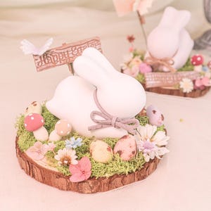 Op de afbeelding: Wit konijnbeeldje op een houten basis met mos, bloemen en paaseieren. Een klein bordje leest "Vrolijk Pasen". Decoratieve paddenstoelen en een vlinder accentueren de scène.