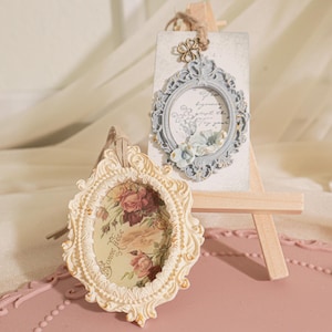 Könnte beinhalten: Zwei verzierte ovale Rahmen mit Blumendesigns. Der Rahmen links ist weiß mit einem Blumenmuster und der Rahmen rechts ist hellblau mit einem Blumenmuster. Beide Rahmen stehen auf Holzstaffeleien.