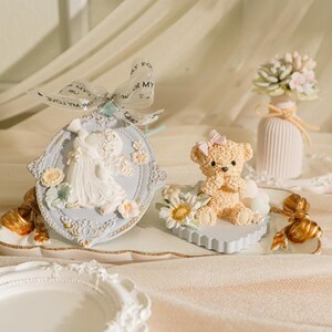 Puede incluir: Una bandeja decorativa con un adorno de cerámica azul y blanco con una pareja, un corazón de cerámica azul claro con un oso de peluche y un jarrón de cerámica blanco con un arreglo floral.