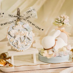 May include: A white ceramic bunny figurine with a brown ribbon around its neck sits on a blue and white base with a white butterfly decoration. The bunny is on a white tray with a gold trim. A white ceramic carousel horse figurine sits on a white bench with a gold trim. The bench is on the same tray as the bunny. The tray is on a white surface. The carousel horse has a blue and white ribbon with the text "MY LOVE", "ONLY FOR MY LOVE", and "FOR MY LOVE" on it.
