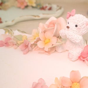 May include: A white, decorative teddy bear figurine with a pink bow sits on a white, floral-decorated tray. The tray is adorned with pink and yellow flowers and green leaves. The overall theme is soft and pastel, with a focus on delicate details.