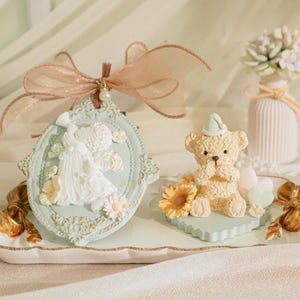 Könnte beinhalten: Ein weißes Porzellantablett mit goldenen Akzenten zeigt ein blaues und weißes Keramikornament mit einer Paarbefigur und einen beigefarbenen Keramikteddybär mit einem blauen Hut. Das Tablett befindet sich auf einer weißen Stoffoberfläche.